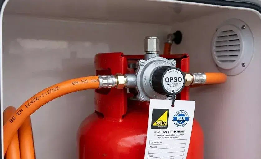 A modern marine LPG regulator with an OPSO safety device and a date-stamped BS EN 16436-1 high-pressure orange hose on a boat.