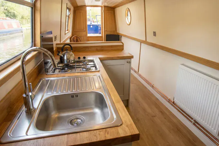 Modern narrowboat galley showing professional LPG gas hob installation, stainless steel plumbing, and a white central heating radiator