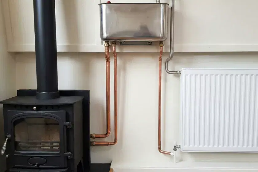 A professional marine back boiler installation featuring simple copper pipework leading to a radiator and a stainless steel open-vent header tank in a narrowboat.