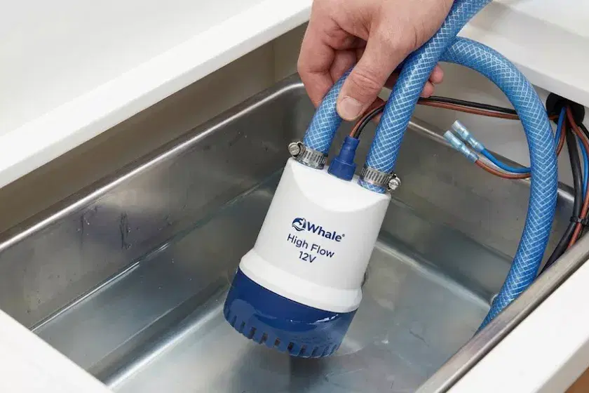 A professional 16:9 photo of a Whale High Flow 12V submersible pump being lowered into a narrowboat freshwater tank with blue reinforced hosing and electrical connections.
