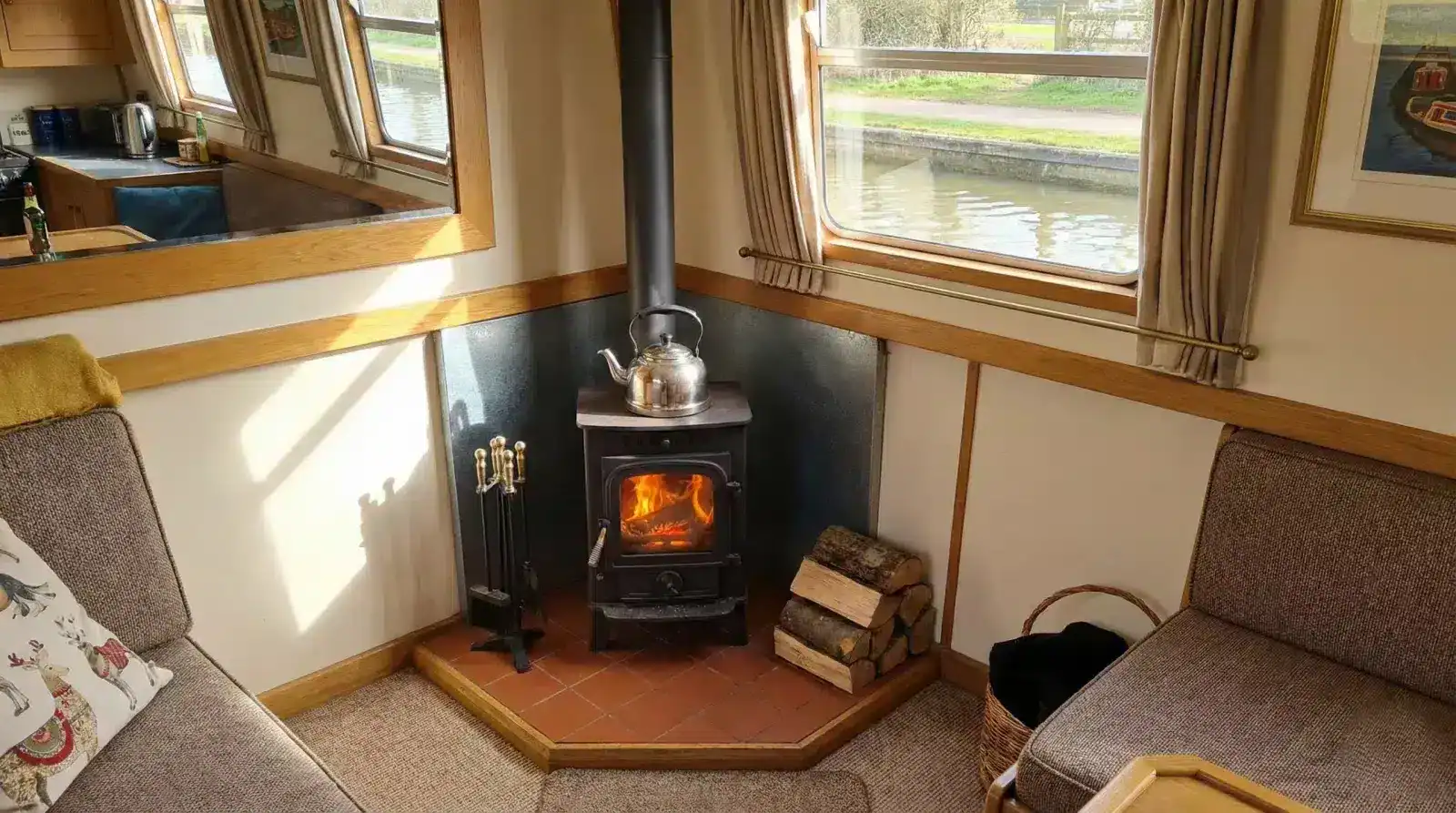 A high-quality interior shot of a narrowboat saloon featuring a professionally installed wood burning stove with stainless steel heat shielding and a view of the UK canal network.