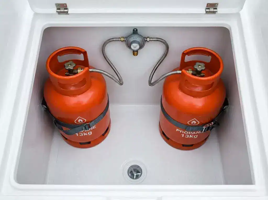 A technical close-up of a compliant boat gas locker showing a central marine-grade changeover regulator with identical stainless steel armored pigtail hoses connected to two 13kg propane cylinders.