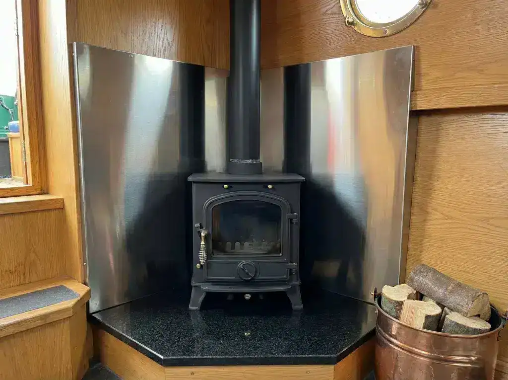 A professionally installed compact marine wood burning stove in a narrowboat corner, featuring stainless steel heat shielding and a tiled hearth compliant with BS 8511 standards.