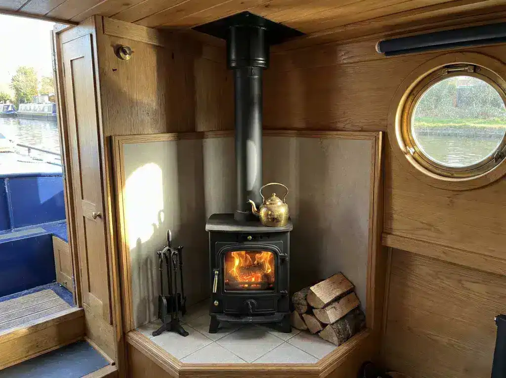 A small black marine wood burning stove installed in the corner of a narrowboat cabin featuring a tiled hearth and professional heat shielding.