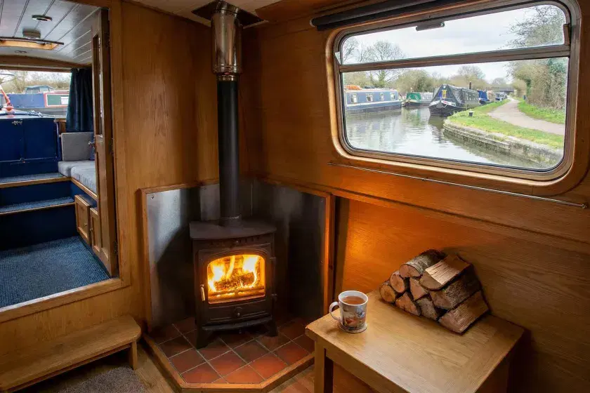 A cozy narrowboat interior featuring a lit wood stove with a clean flue installation and views of a UK canal through the window.