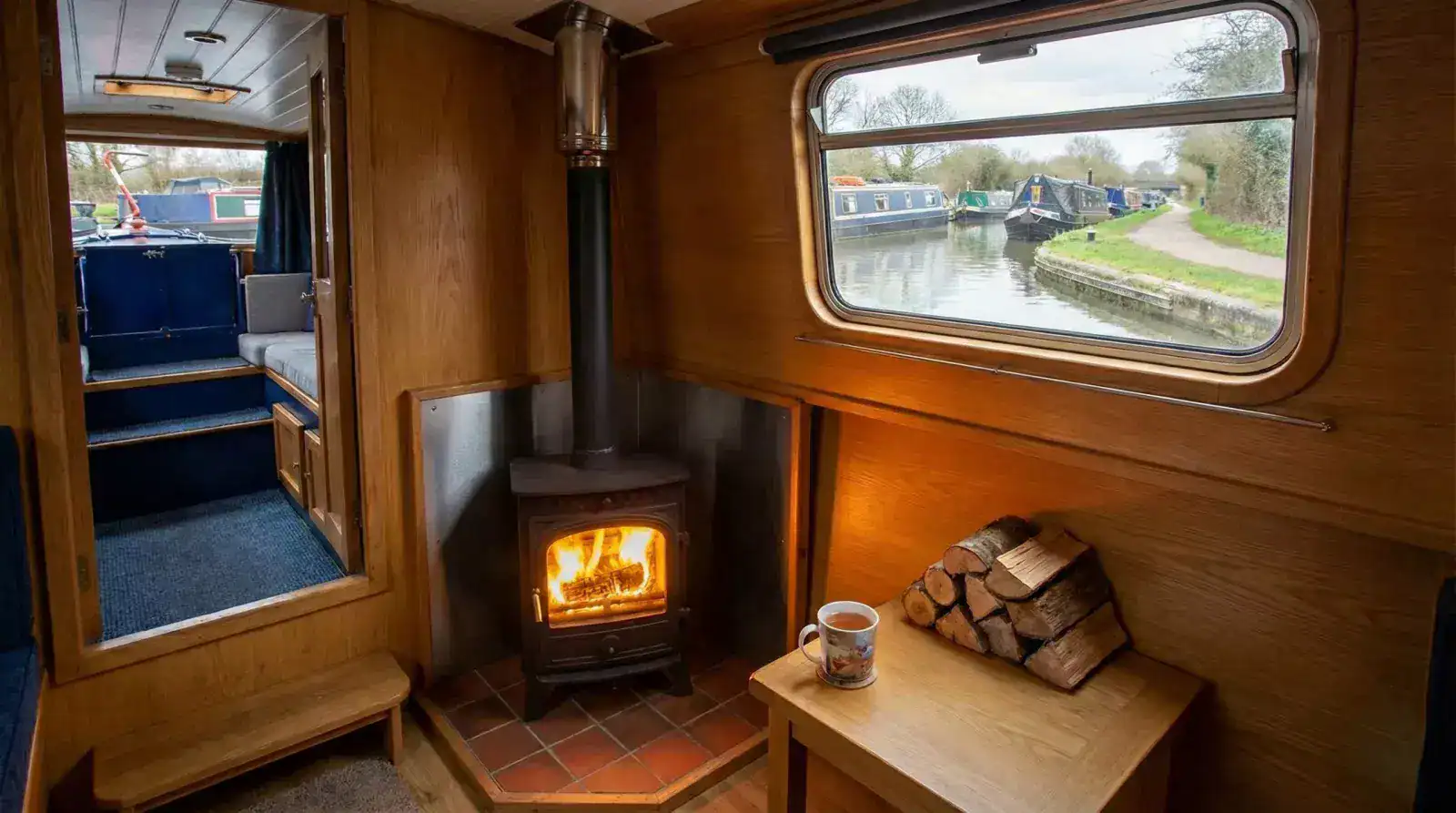 A cozy narrowboat interior featuring a lit wood stove with a clean flue installation and views of a UK canal through the window.