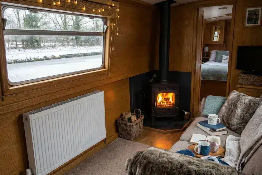 A warm and cozy narrowboat interior during winter, showing a glowing wood burning stove and a wall-mounted radiator for dual heating.