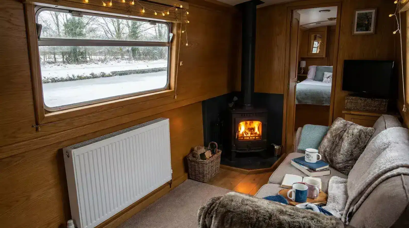 A warm and cozy narrowboat interior during winter, showing a glowing wood burning stove and a wall-mounted radiator for dual heating.