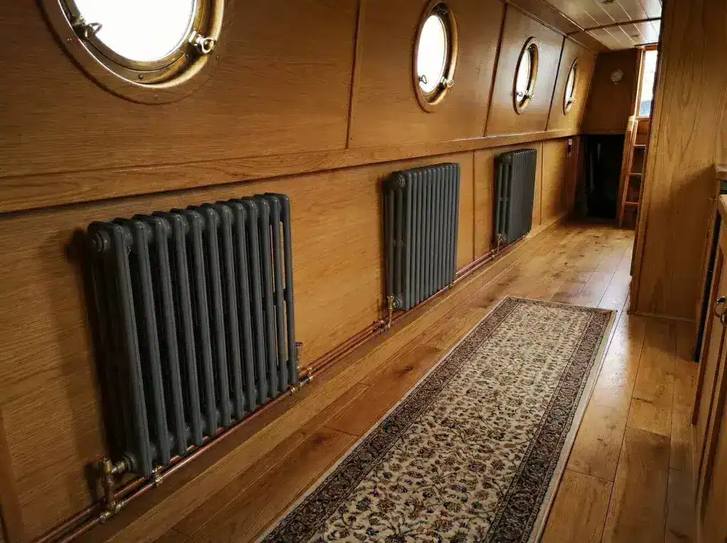 A row of traditional-style dark grey radiators installed along a wood-panelled narrowboat corridor for central heating.