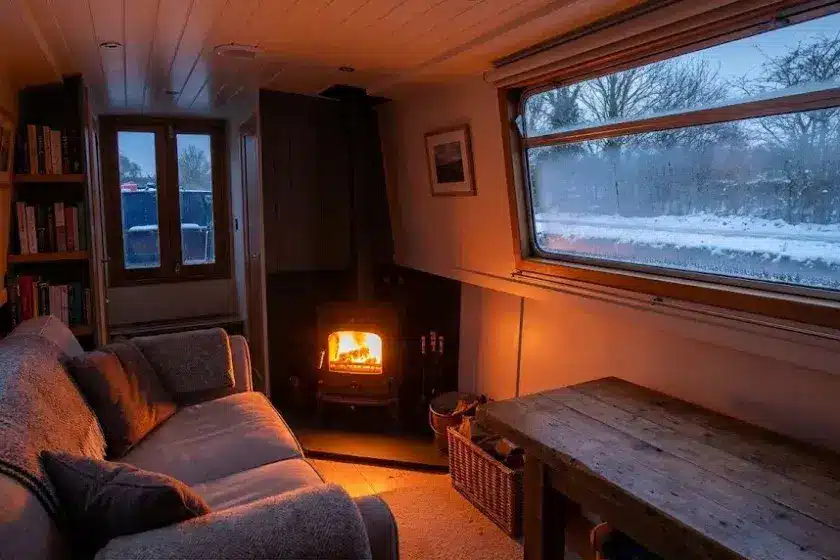 A cozy narrowboat interior during winter featuring a lit solid fuel stove and a snowy canal towpath visible through the window.