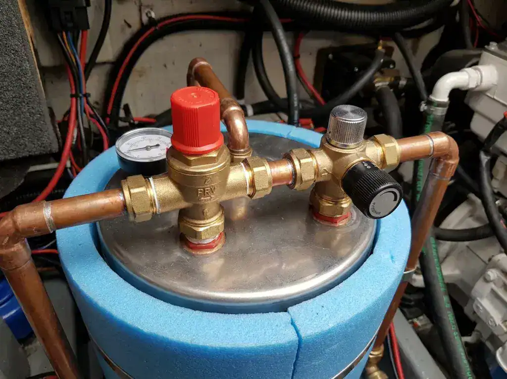 A technical close-up of a boat calorifier showing the pressure release valve (PRV) and a thermostatic mixing valve with brass plumbing fittings.