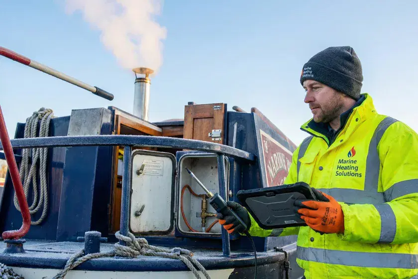 A professional Marine Heating Solutions technician in branded gear standing on a narrowboat stern with a digital tablet during a winter LPG safety inspection.
