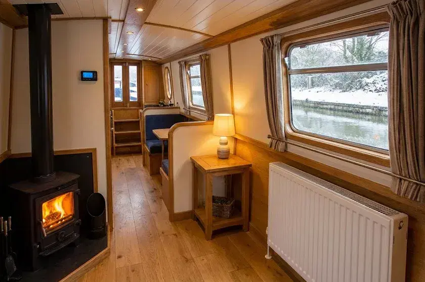 A cozy narrowboat interior in winter showing a complete central heating system with a solid fuel stove, a modern white radiator, and a digital thermostat.