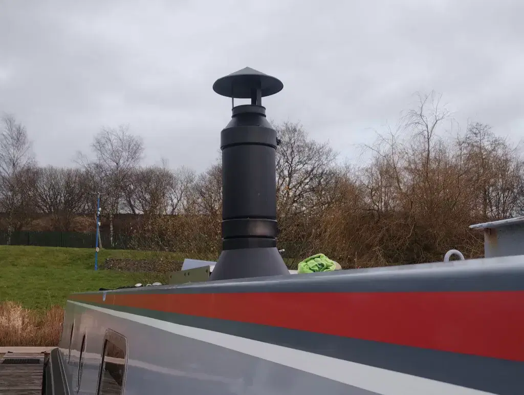 A close-up of a black powder-coated twin-wall insulated flue kit installed on a narrowboat roof, showing the rain cowl and secure roof mounting.
