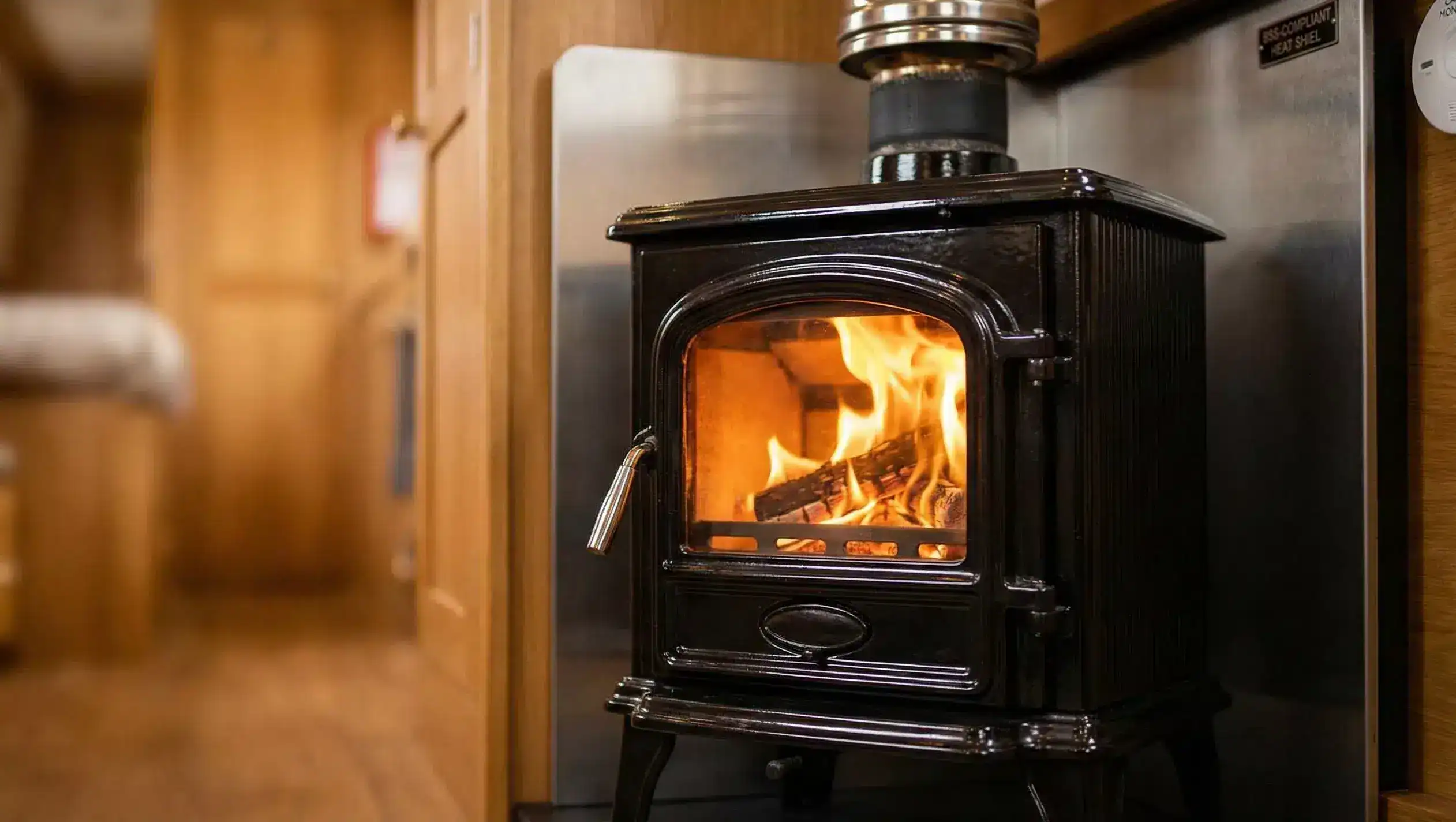 A professionally installed solid fuel boat stove with a fire-lit chamber, twin-wall insulated flue, and BSS-compliant heat shielding in a narrowboat interior.