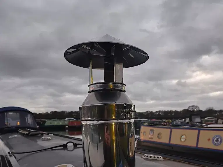 A close-up of a 316L stainless steel twin-wall flue assembly, including the rain cowl and locking band, installed on a narrowboat roof at a marina.
