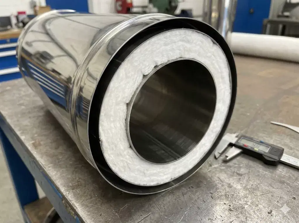 A technical close-up of a cutaway twin-wall flue section, showing the high-density insulation layer sandwiched between the stainless steel inner and outer pipes.