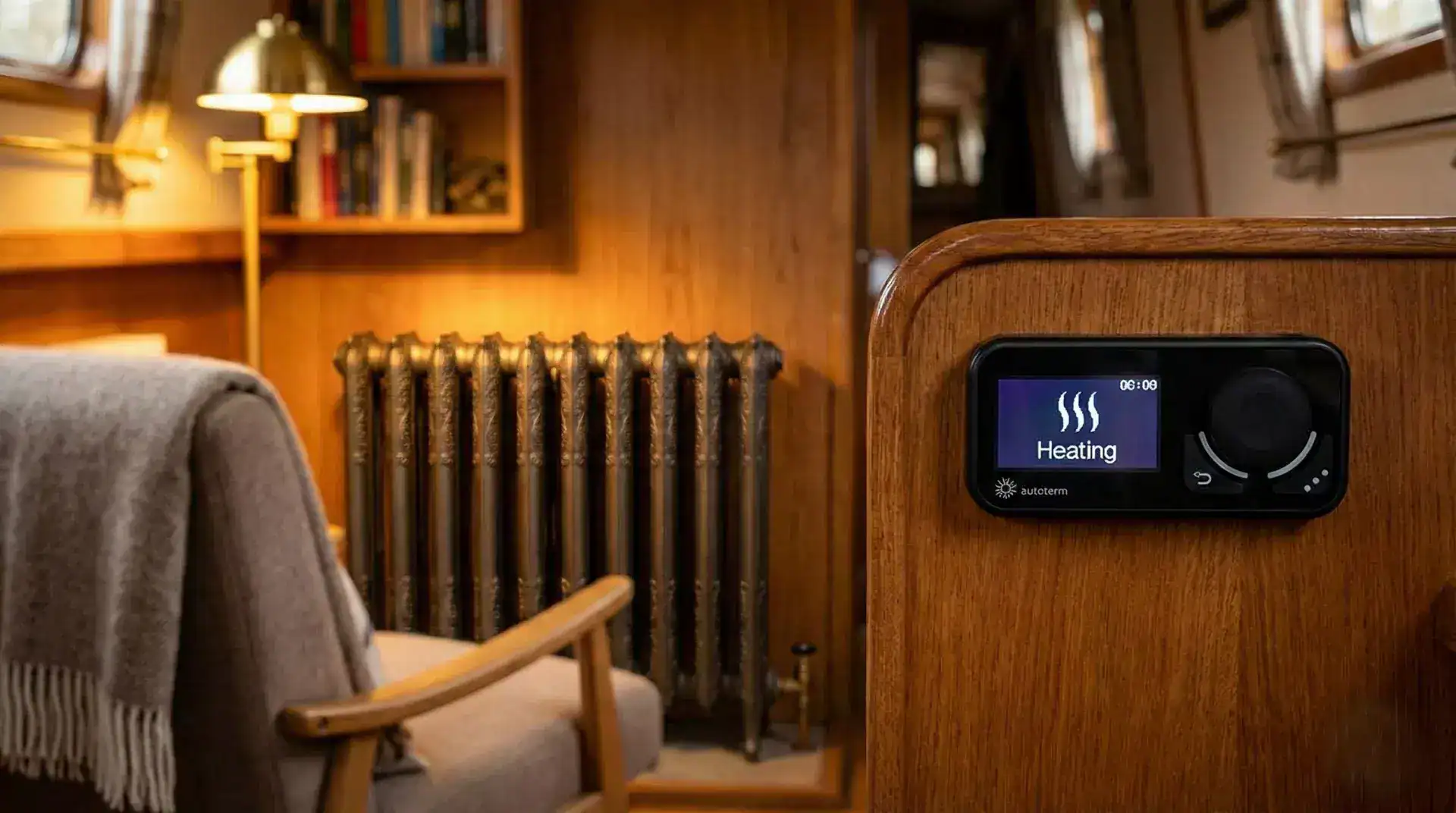 An Autoterm Comfort Control digital thermostat mounted on a wooden wall in a warm boat cabin, with a traditional cast iron radiator in the background.
