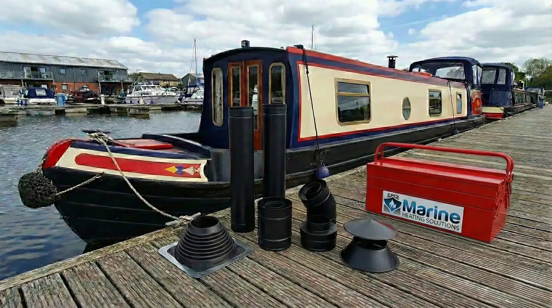 Black powder-coated twin wall insulated flue kit with a black roof flashing collar displayed on a wooden pontoon next to a narrowboat, featuring a red Marine Heating Solutions toolbox.