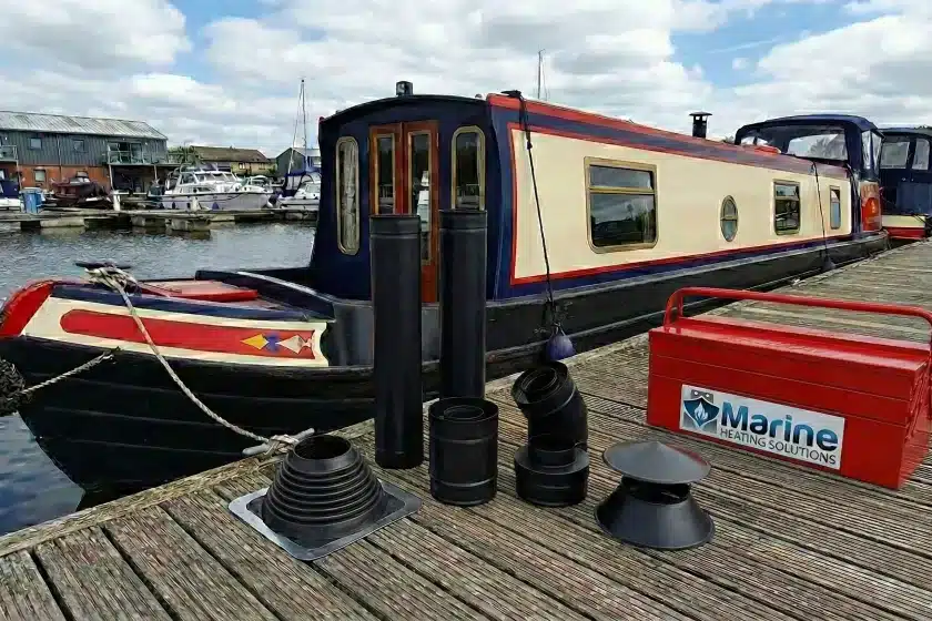 Black powder-coated twin wall insulated flue kit with a black roof flashing collar displayed on a wooden pontoon next to a narrowboat, featuring a red Marine Heating Solutions toolbox.