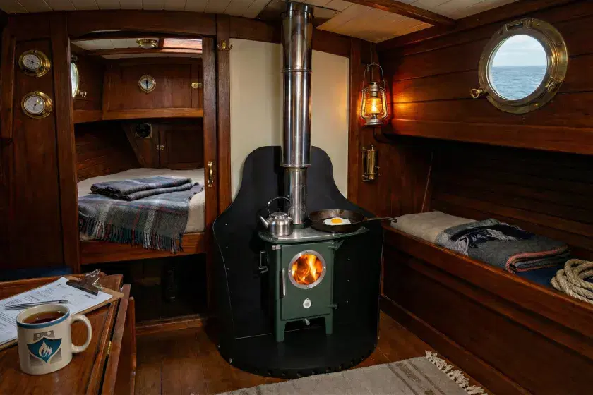 A green Chilli Billie Gentoo wood-burning stove with a circular glass door and active fire, professionally installed in a wooden boat cabin by Marine Heating Solutions.