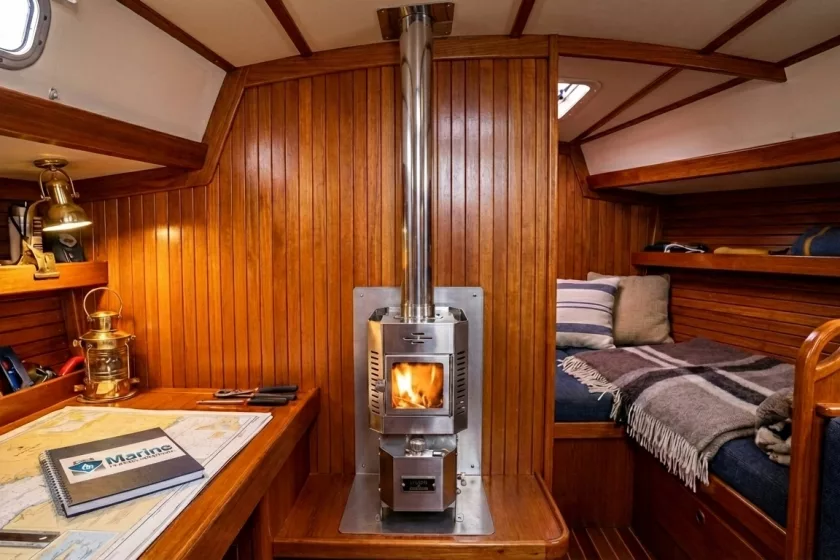 A Dickinson Alaska floor-mounted diesel heater with a vertical stainless steel flue installed in a traditional wood-paneled boat cabin, featuring a Marine Heating Solutions notebook on the chart table.