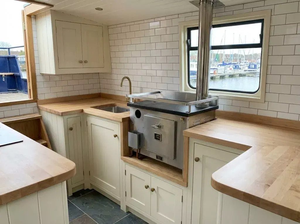 A Dickinson Bering diesel cookstove professionally installed on a wooden plinth within a modern narrowboat galley, featuring white subway tiles and a stainless steel flue.