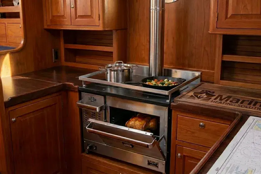 A Dickinson Pacific diesel cooker installed in a traditional boat galley featuring wood cabinetry, a stainless steel flue, and fresh food cooking in the oven and on the cooktop.
