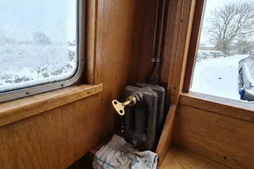 A technical close-up of a brass bleed key inserted into a narrowboat radiator valve, showing a snowy canal scene through the window to emphasize winter maintenance.