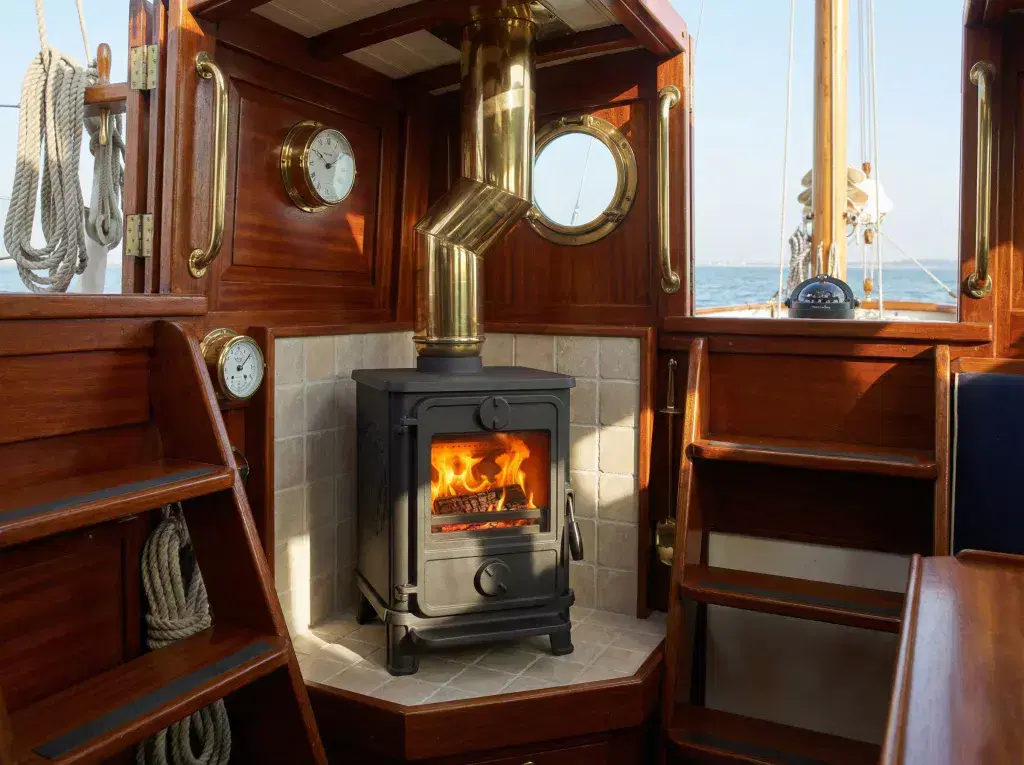 A black Morso Squirrel 1412 multi-fuel stove with a lit fire, installed on a beige tiled hearth inside a classic wooden yacht cabin featuring brass fixtures and a view of the sea.