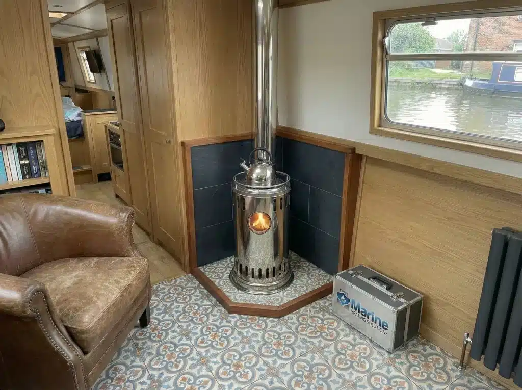 A stainless steel Refleks 62M diesel boat stove with a viewing window and kettle, installed in a cozy narrowboat interior with a tiled hearth.