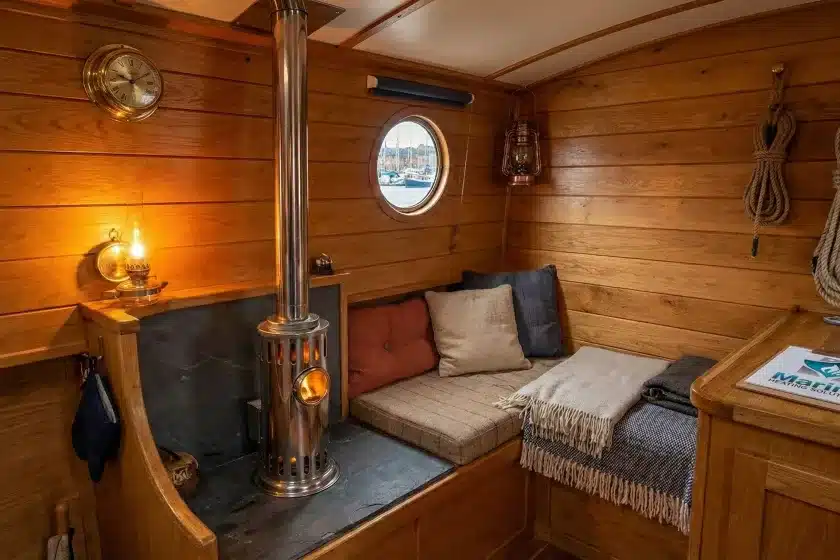 A stainless steel Refleks 66M diesel boat stove installed in a cozy, wood-paneled boat cabin, featuring a clipboard with the Marine Heating Solutions logo on a wooden sideboard.