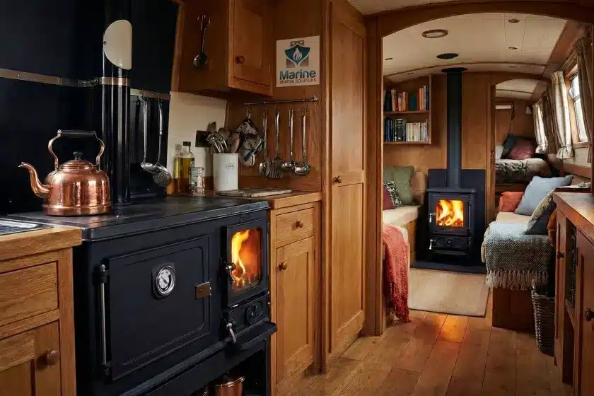 A cozy narrowboat interior featuring a Salamander Little Range multi-fuel cooker in the galley foreground and a Salamander Hobbit stove in the lounge background.