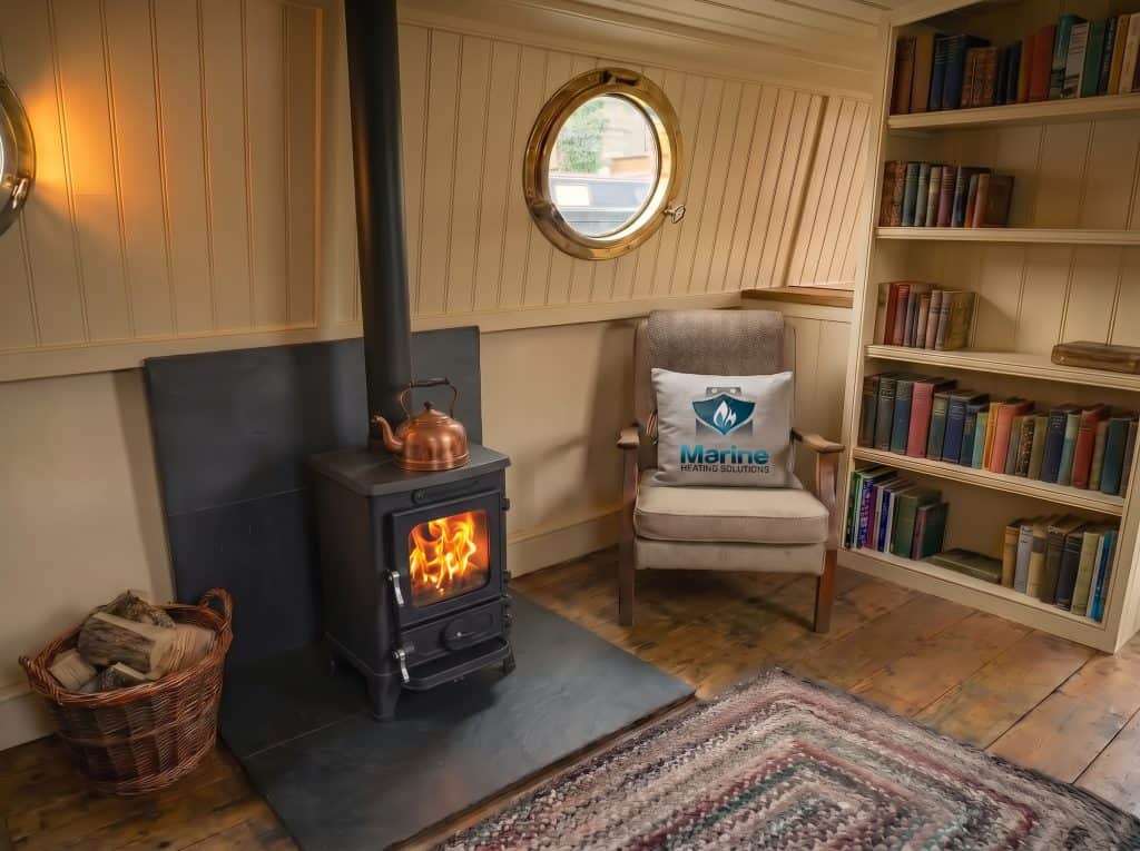 A cozy narrowboat interior featuring a lit Salamander Hobbit stove, a Marine Heating Solutions branded toolbox on the floor, and a gray cushion with the company logo on a wooden armchair.