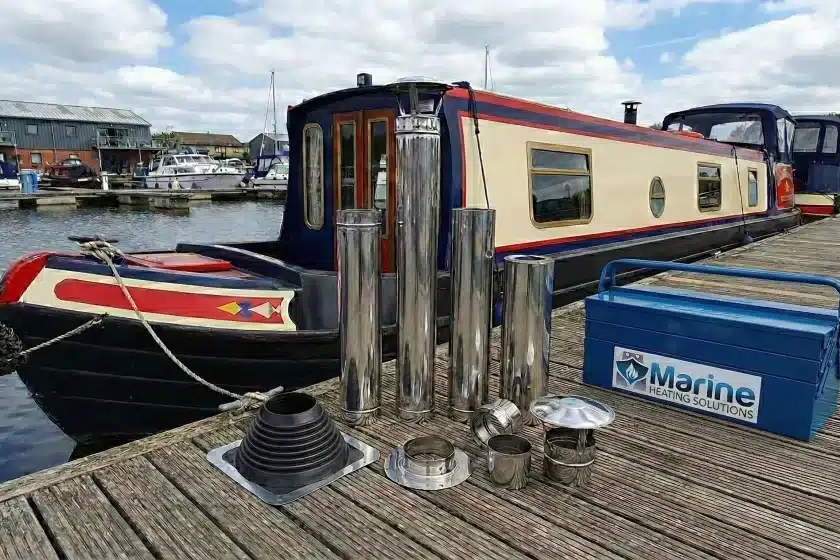 Stainless steel twin wall insulated flue kit with a black roof flashing collar displayed on a wooden pontoon next to a narrowboat.