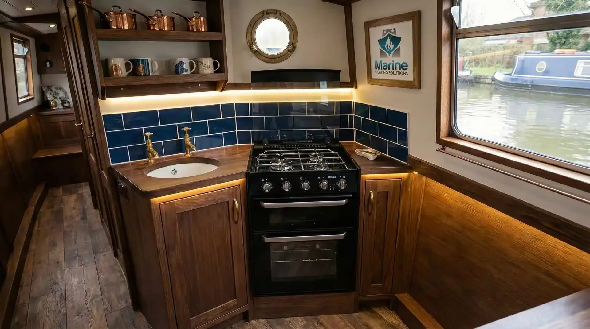 A black Thetford Caprice 3 LPG cooker installed in a modern narrowboat kitchen with wood cabinetry and blue tile backsplash, featuring a picture frame with the Marine Heating Solutions logo on the wall.