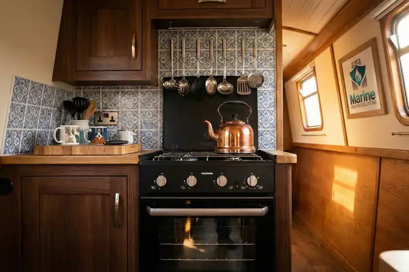 A Thetford Triplex black LPG 3-in-1 cooker and oven installed in a traditional narrowboat kitchen featuring wood cabinetry and blue patterned tiles.