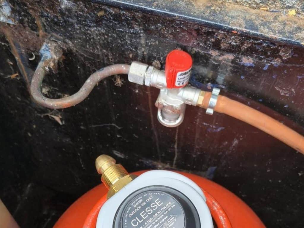 An Alde bubble tester gas leak detector with a red bypass button mounted in a boat gas locker, connected to copper piping and an orange LPG hose above a Clesse propane regulator.