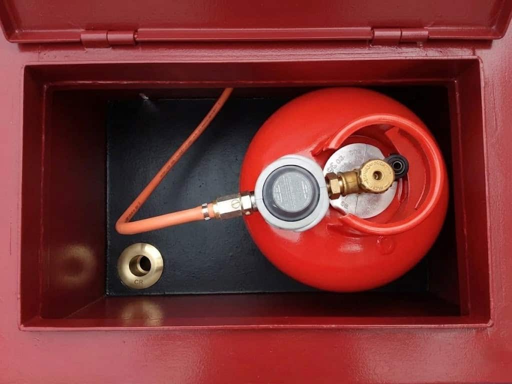 Top-down view of a compliant boat gas locker showing a clean red LPG cylinder, gas regulator, orange flexible hose, and an unobstructed brass low-level drain hole.
