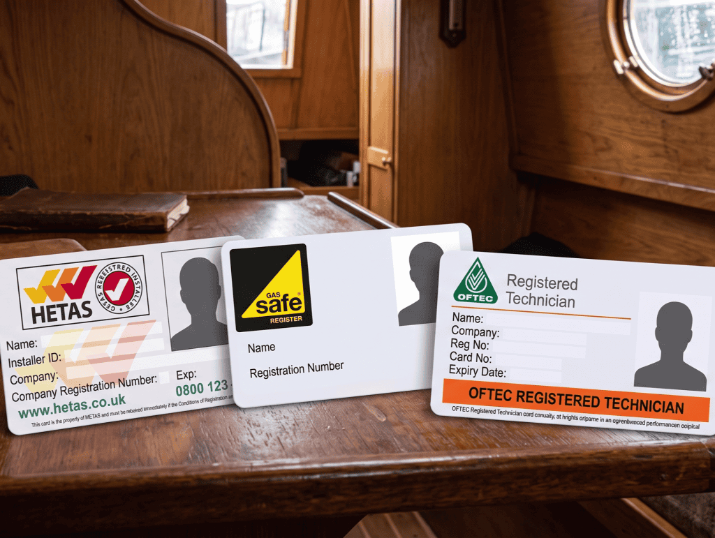 Three professional trade qualification ID cards for HETAS, Gas Safe Register, and OFTEC displayed on a wooden table inside a boat cabin.
