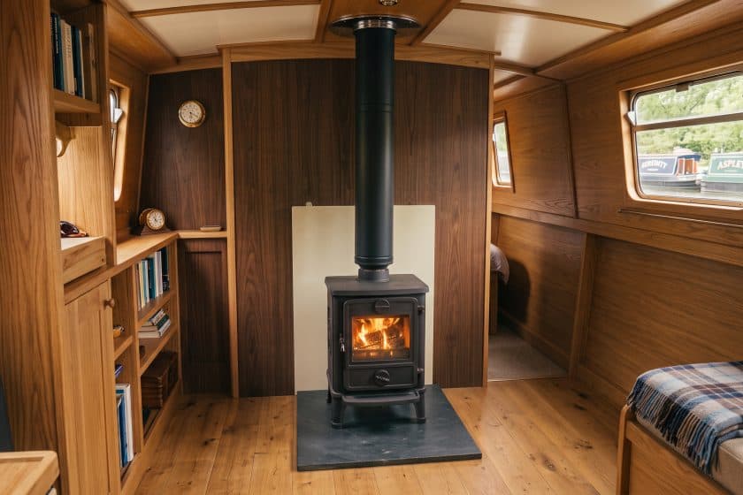 Professional solid fuel stove installation on a narrowboat featuring a matte-black twin-wall insulated flue, slate hearth, and air-gapped thermal heat shield.