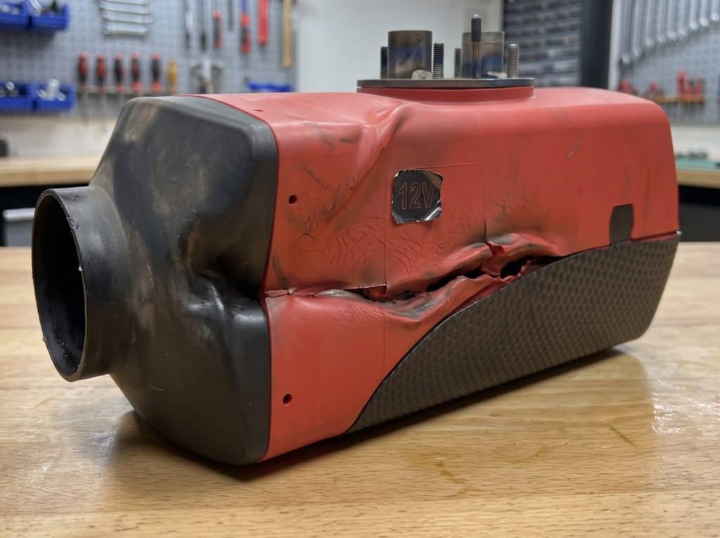 A close-up photograph of a red and black portable diesel air heater with its plastic casing severely warped, melted, cracked, and discolored due to extreme overheating, sitting on a wooden workbench.